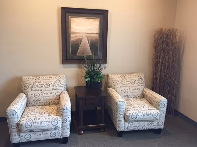 Two patterned armchairs flank a dark wooden side table with a potted plant beneath a framed pier print and a bundle of decorative dried branches in a neutral seating area.