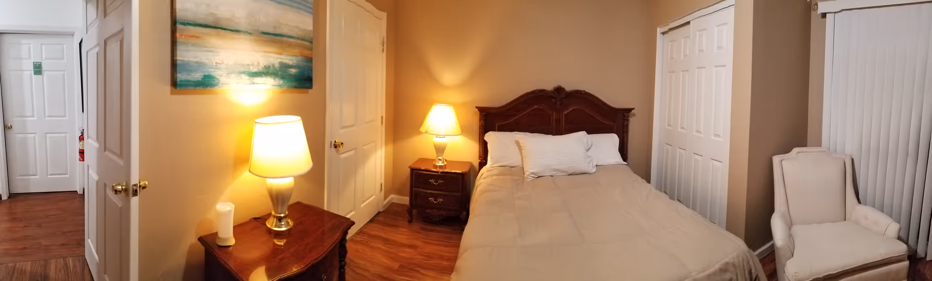 A cozy bedroom with a wooden bed frame, beige bedding, and two white pillows. There are two wooden nightstands on either side of the bed, each with a lit table lamp. To the left, there is a wooden dresser with a lamp and a candle on it, and above the dresser hangs a colorful abstract painting. The room has beige walls, wood flooring, a white armchair near a window with vertical blinds, and multiple white doors including a closet and an entrance door.