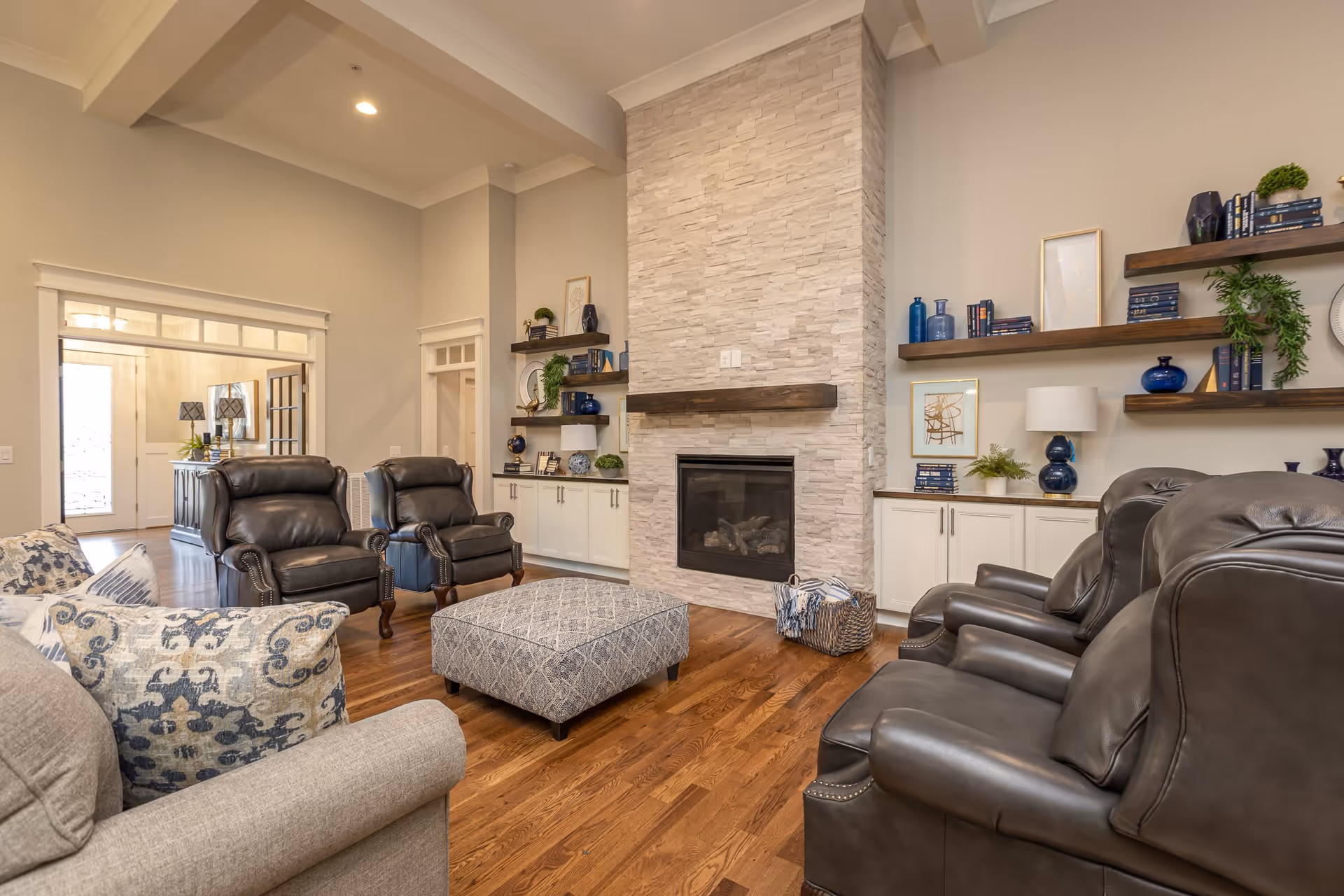 A cozy living room with a stone fireplace in the center, flanked by built-in white cabinets and wooden shelves decorated with books, plants, and vases. The room features dark leather armchairs, a patterned ottoman, and a beige sofa with decorative pillows. Hardwood floors and soft lighting create a warm atmosphere.