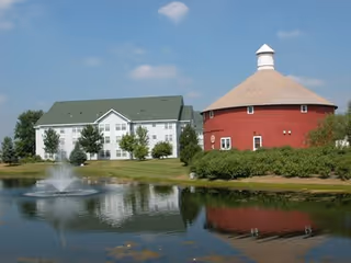 A senior living facility with a white multi-story building and a large red round barn-like structure next to a pond with a water fountain, surrounded by green grass and trees under a blue sky with some clouds.