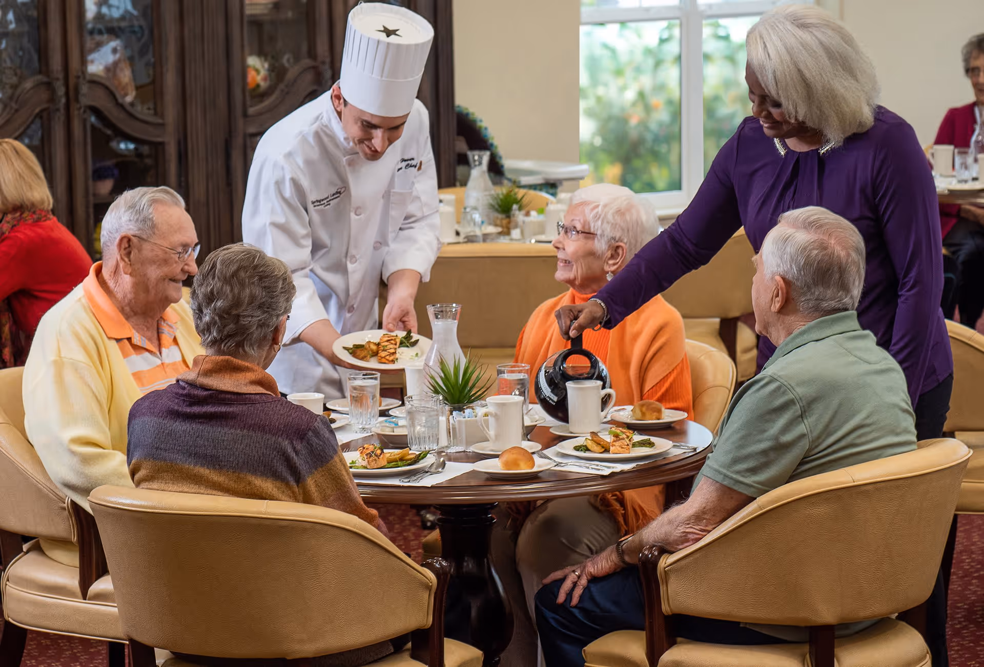 A group of four elderly people sitting around a dining table in a retirement facility, being served food by a chef in a white uniform and a woman pouring coffee. The table is set with plates of food, glasses of water, and coffee cups. The setting is warm and inviting with large windows in the background.