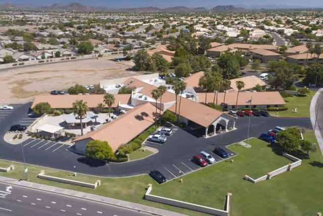 Aerial view of Lake Pleasant Post Acute Rehabilitation Center showing a single-story building with a terracotta tiled roof, surrounded by parking lots with several cars, palm trees, and green lawns. The facility is located in a suburban area with many houses and mountains visible in the background.