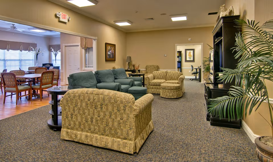 Carpeted senior living common room with upholstered sofas and chairs, TV cabinet, plants, and a dining area visible through an open doorway.
