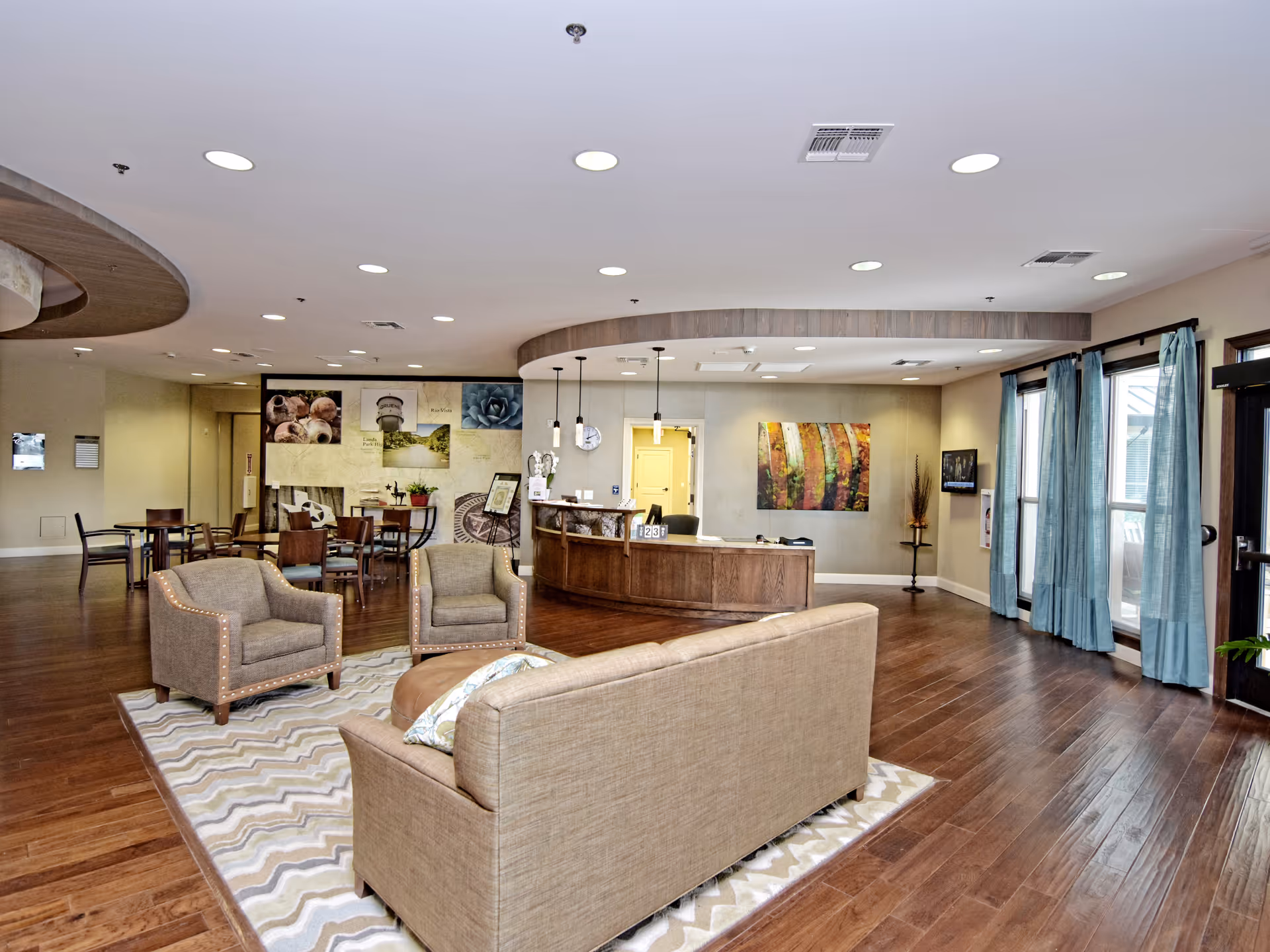 Open communal living area with sofas and armchairs on a rug, a reception desk and dining tables in the background.