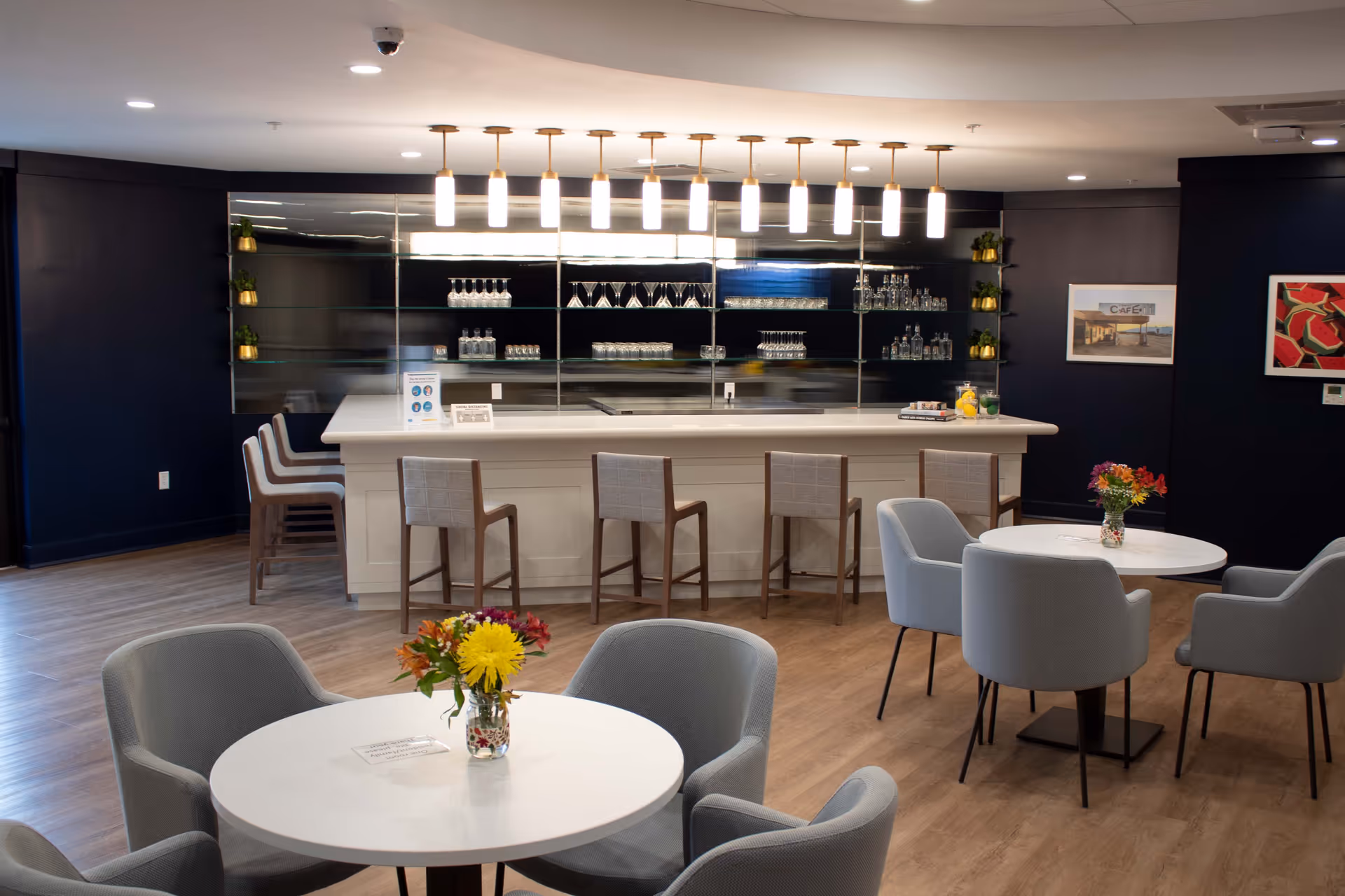 A modern indoor lounge area with a white bar counter and five bar stools. Behind the bar is a mirrored wall with shelves holding glassware and bottles. In the foreground, there are round white tables with gray upholstered chairs, each table decorated with a small vase of colorful flowers. The room has wooden flooring and dark blue walls with framed artwork.