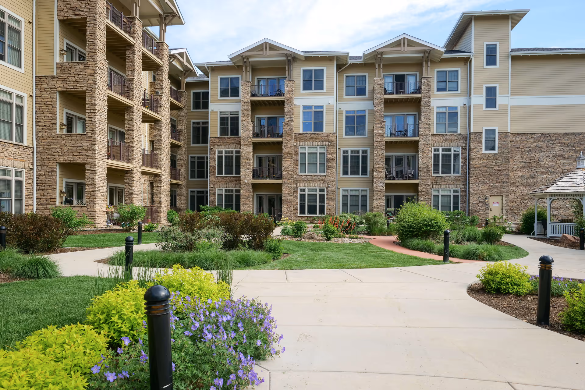 Exterior view of a multi-story senior living facility building with stone and beige siding, balconies, and numerous windows. In the foreground, there is a well-maintained garden with green shrubs, purple flowers, and paved walkways leading to the building. A white gazebo is visible on the right side.