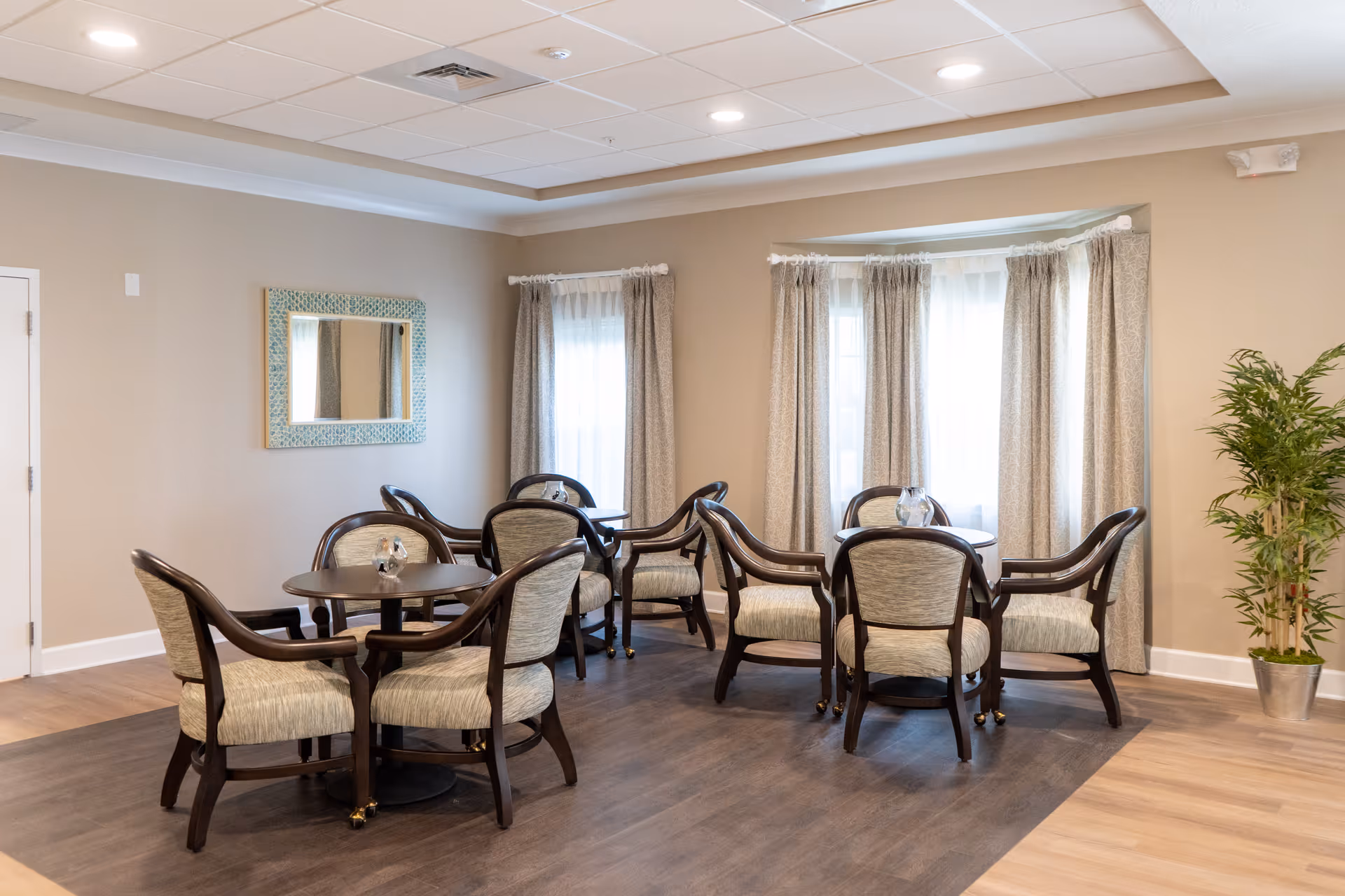 A cozy interior room with two round tables, each surrounded by four cushioned chairs with wooden frames. The room has light beige walls, a large window with curtains allowing natural light, a decorative mirror on one wall, and a potted plant in the corner. The floor has a combination of wood and carpeted areas.