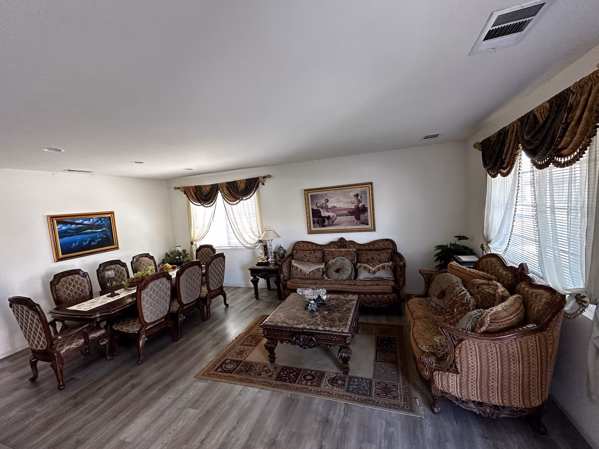 A spacious living and dining area with classic wooden furniture. The living room features two ornate sofas with patterned cushions, a wooden coffee table on a decorative rug, and framed artwork on the walls. The dining area has a long wooden table with eight upholstered chairs. Large windows with sheer curtains and valances allow natural light to fill the room. The floor is covered with light-colored wood laminate.