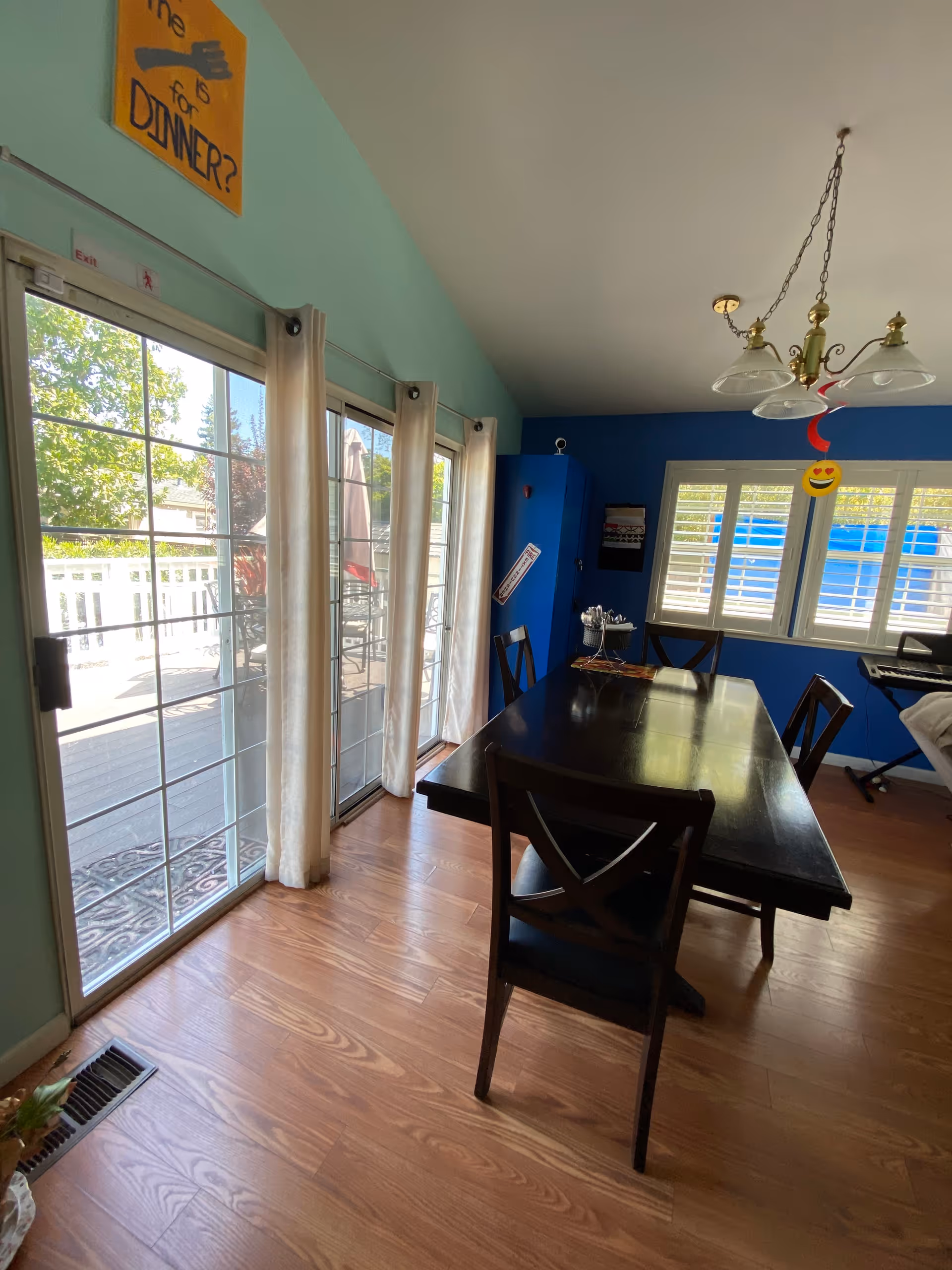 Dining room with a dark wooden table and four chairs on a wooden floor. Large sliding glass doors with white curtains lead to an outdoor patio. The walls are painted light green and blue, with a window featuring white shutters. A chandelier with three glass shades hangs above the table. A yellow sign on the wall reads 'Time is for DINNER?'.