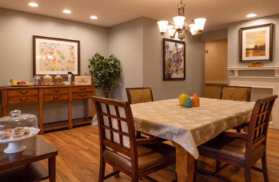 A cozy dining room with a wooden table covered by a beige floral tablecloth, surrounded by four wooden chairs with cushioned seats. On the table are three colorful ceramic jars. The room features warm wooden flooring, a wooden sideboard with jars and fruit, framed artwork on the walls, a potted plant in the corner, and a chandelier with multiple lights hanging from the ceiling.