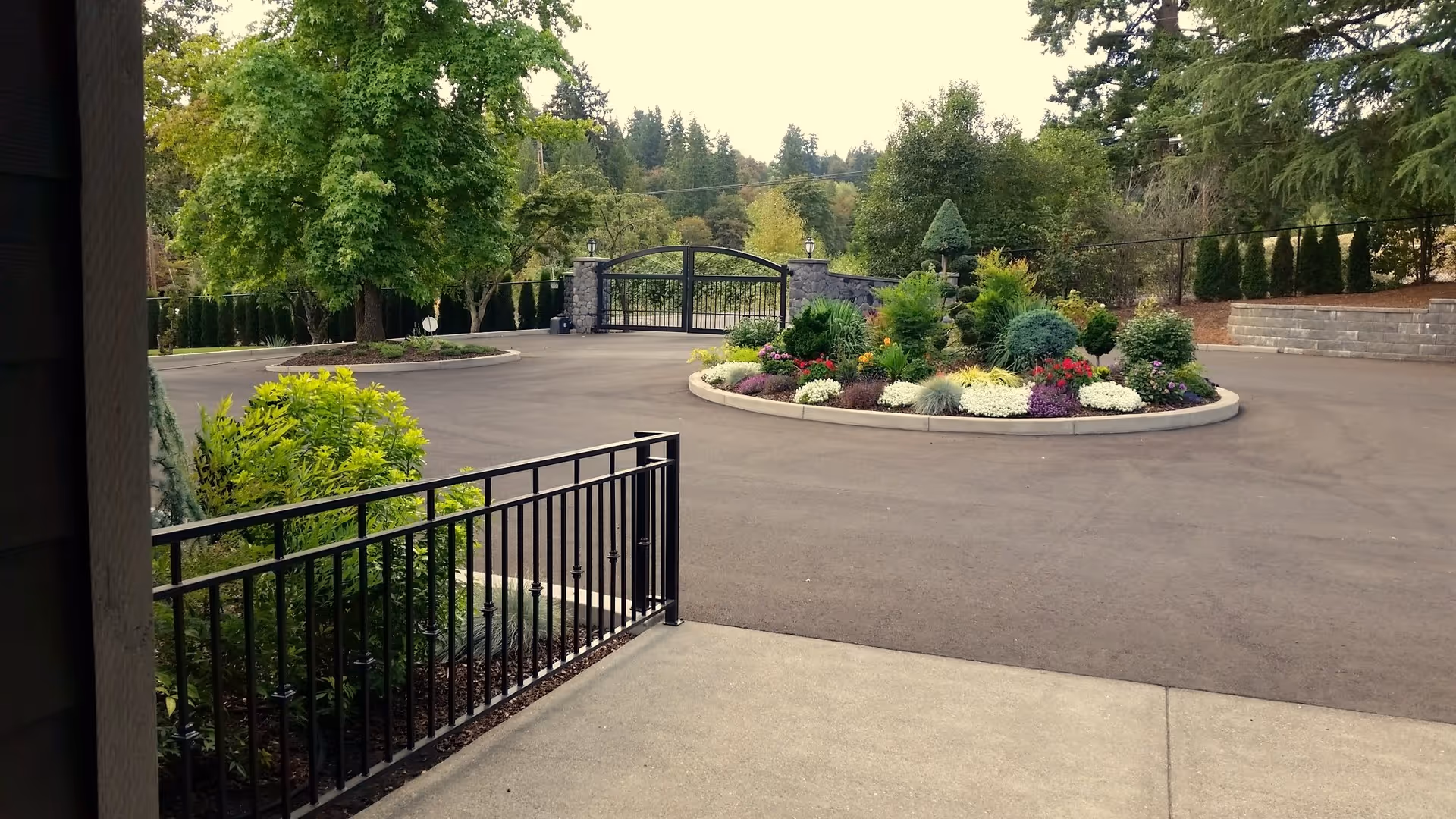 View of a paved driveway with a circular flower bed in the center, featuring various colorful flowers and shrubs. There is a black metal gate and stone pillars at the end of the driveway, surrounded by tall trees and greenery.