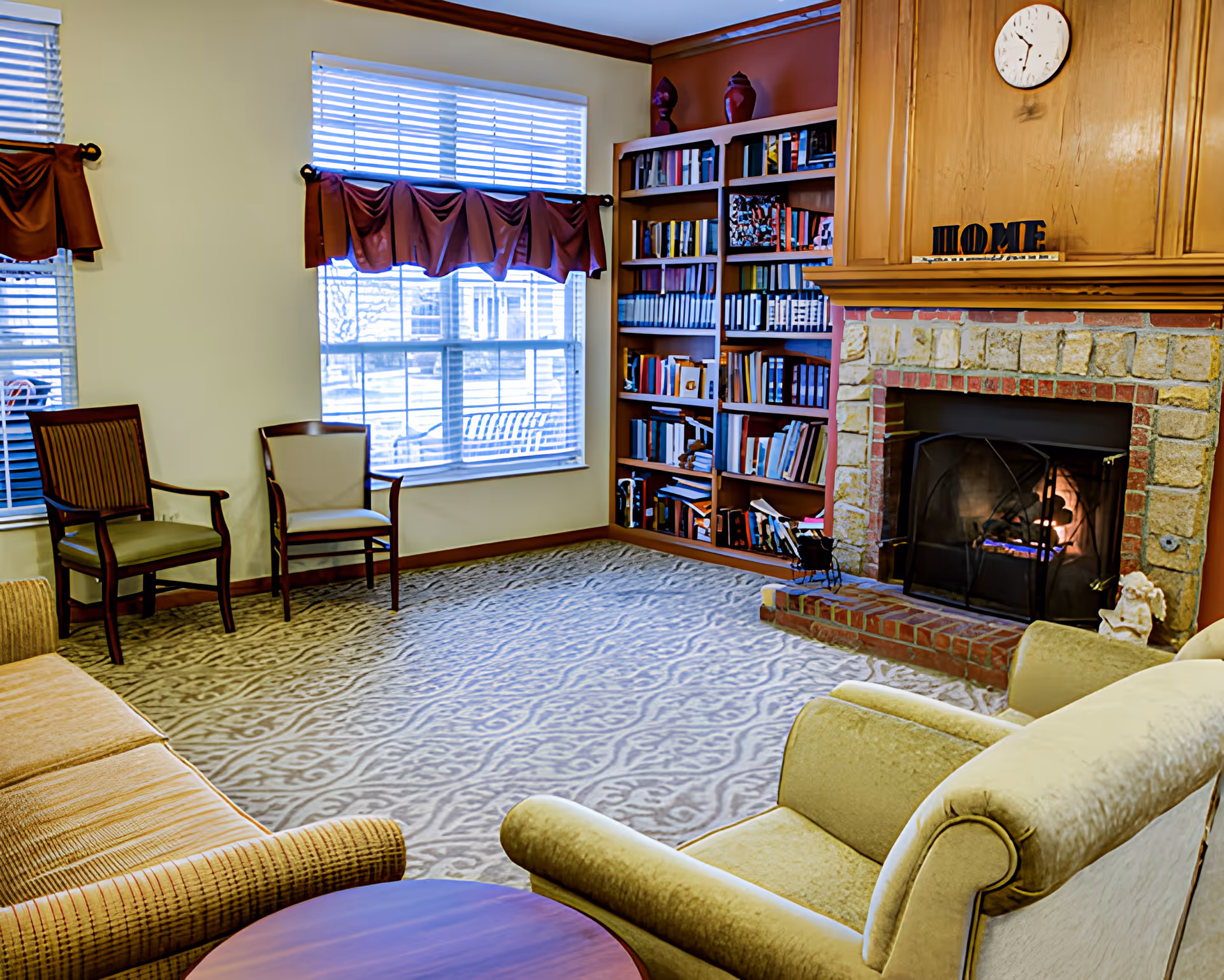 Cozy living room with armchairs and a sofa, a lit brick fireplace, built-in bookshelves, and large windows with valances.