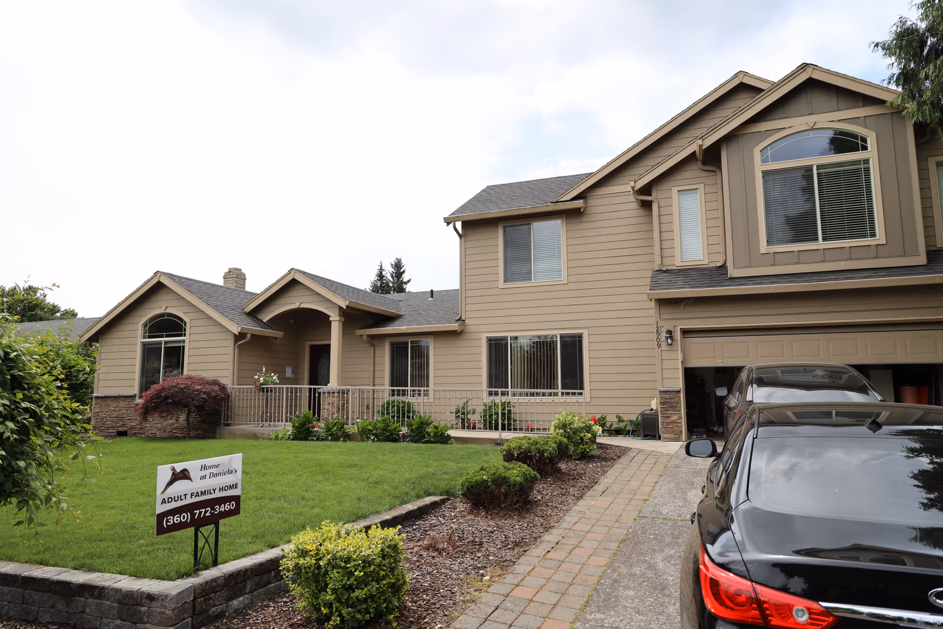 Exterior view of a two-story beige house with a well-maintained front yard, a driveway with two parked cars, and a sign in the lawn that reads 'Home at Daniela's Adult Family Home (360) 772-3460'.