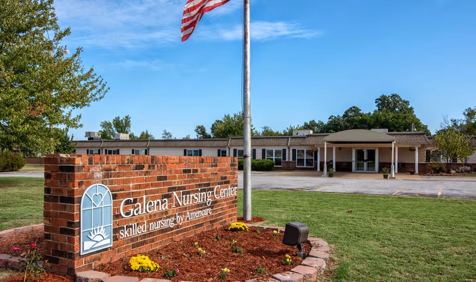 Exterior view of Galena Nursing Center showing a brick sign with the facility name and logo, an American flag on a flagpole, a well-maintained lawn with flowers, and the entrance to the single-story building under a clear blue sky.