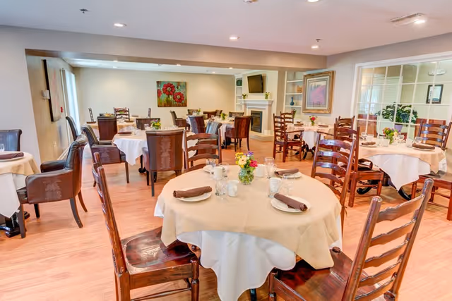 A bright and inviting dining room with multiple round tables covered with white tablecloths and beige overlays. Each table is set with plates, glasses, napkins, and small flower arrangements. The room features wooden chairs, a fireplace with a TV mounted above it, and artwork on the walls. Large windows and glass panels allow natural light to fill the space.