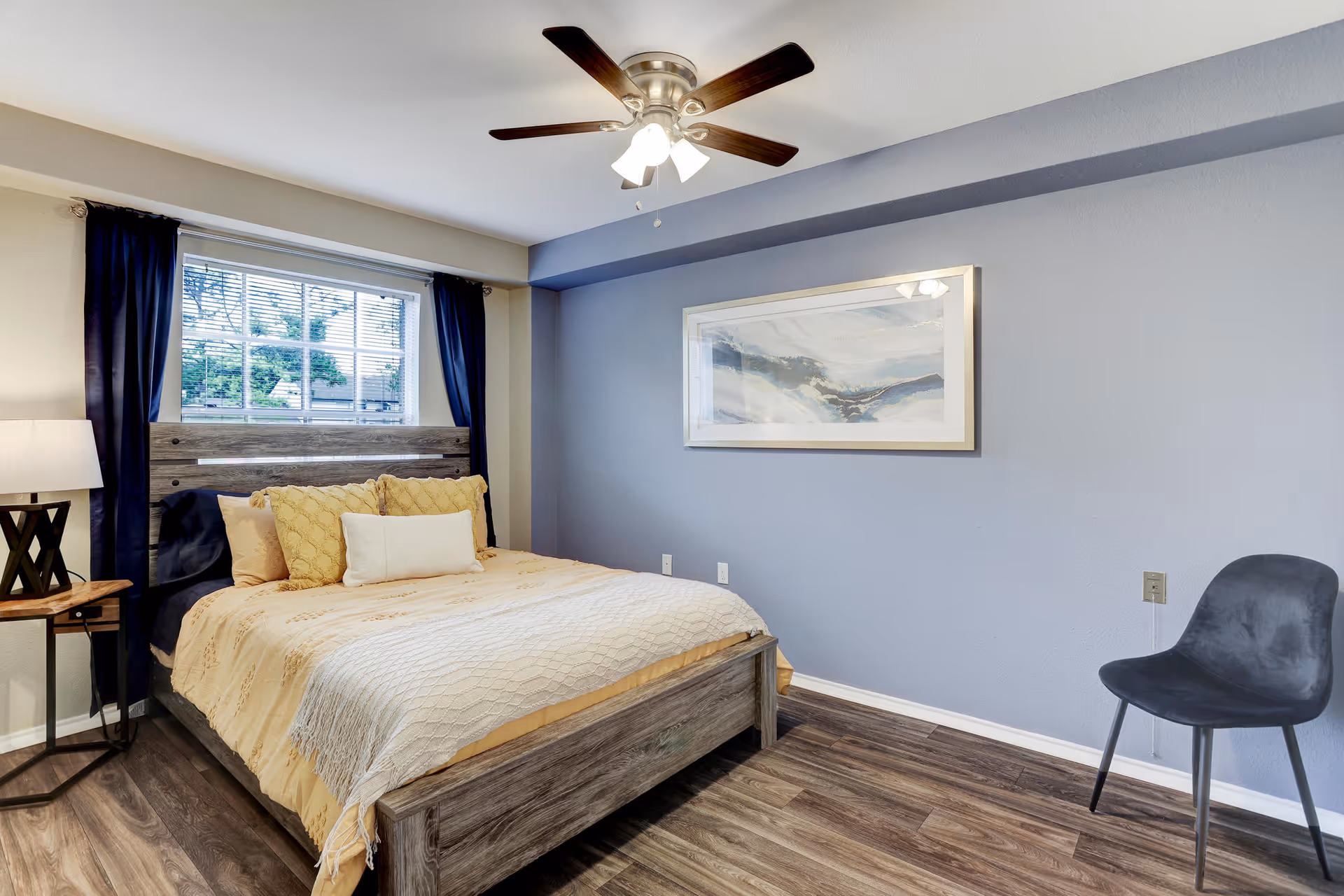 A bedroom with a wooden bed frame, beige bedding, and multiple pillows. There is a window with dark curtains behind the bed, a wooden side table with a lamp on the left, a ceiling fan with lights above, a framed abstract painting on the light blue wall, and a dark gray chair on the right side of the room.