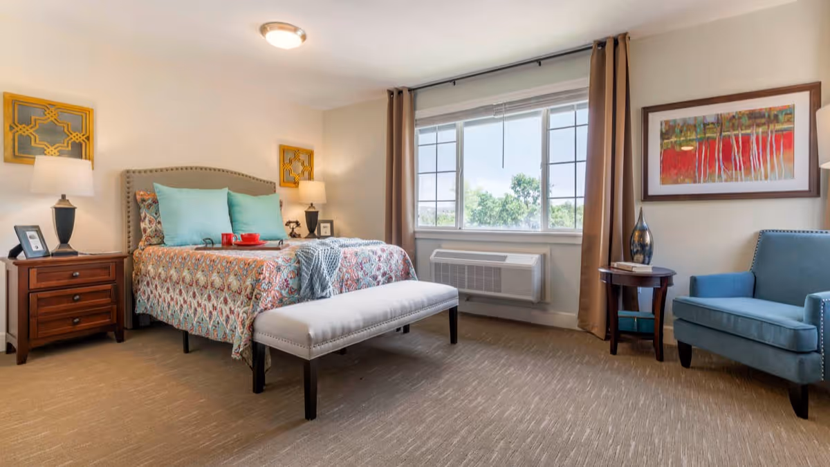 A well-lit bedroom with a large bed featuring patterned bedding and turquoise pillows. There are two bedside tables with lamps and decorative items. A cushioned bench is placed at the foot of the bed. A large window with brown curtains lets in natural light, and a blue armchair and small round table with a vase and books are positioned near the window. A colorful framed painting hangs on the wall.