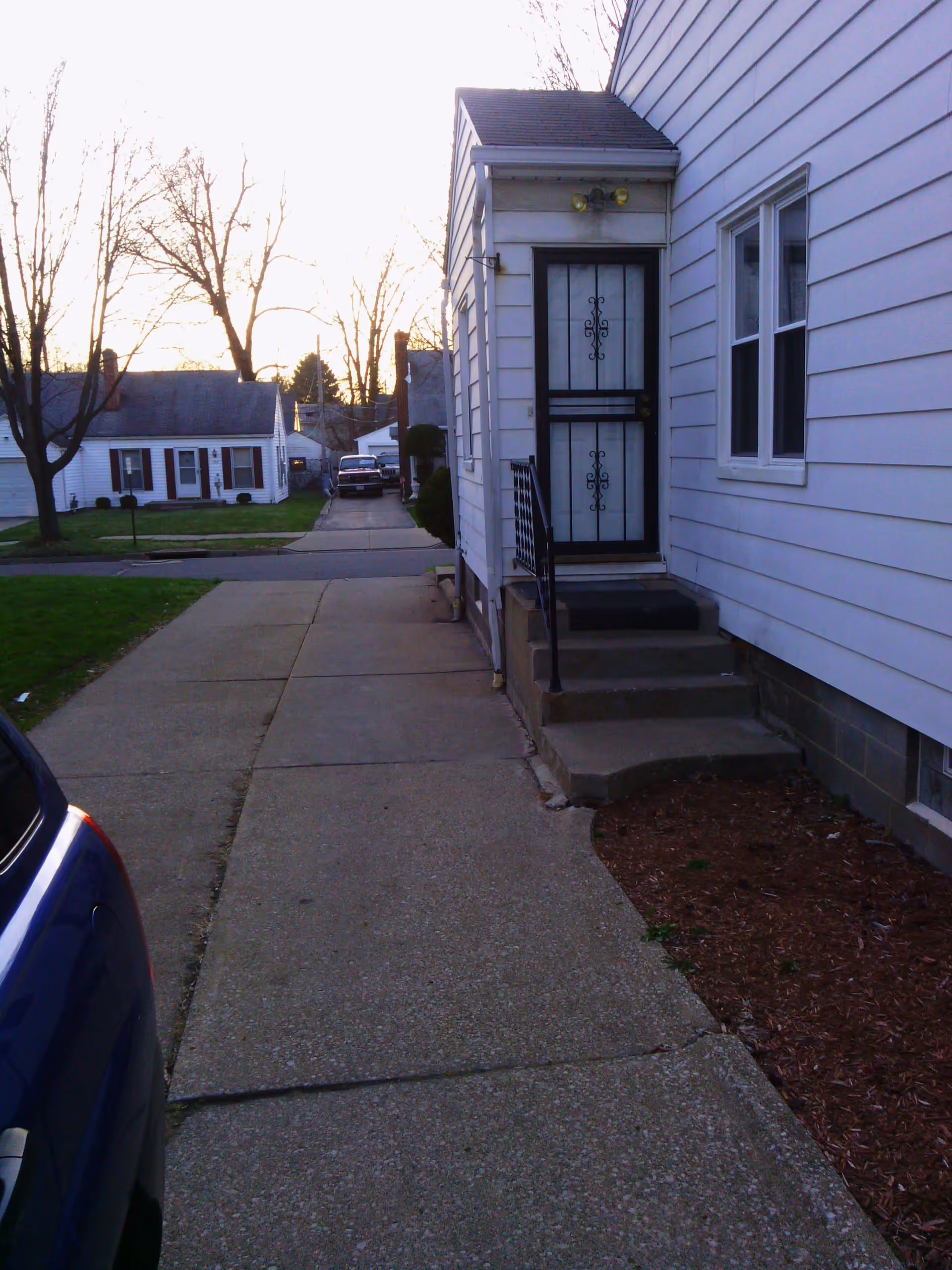 Sidewalk leading to the entrance of a white house with a black metal security door and small steps. There is a blue car partially visible on the left side and other houses and trees in the background during sunset.