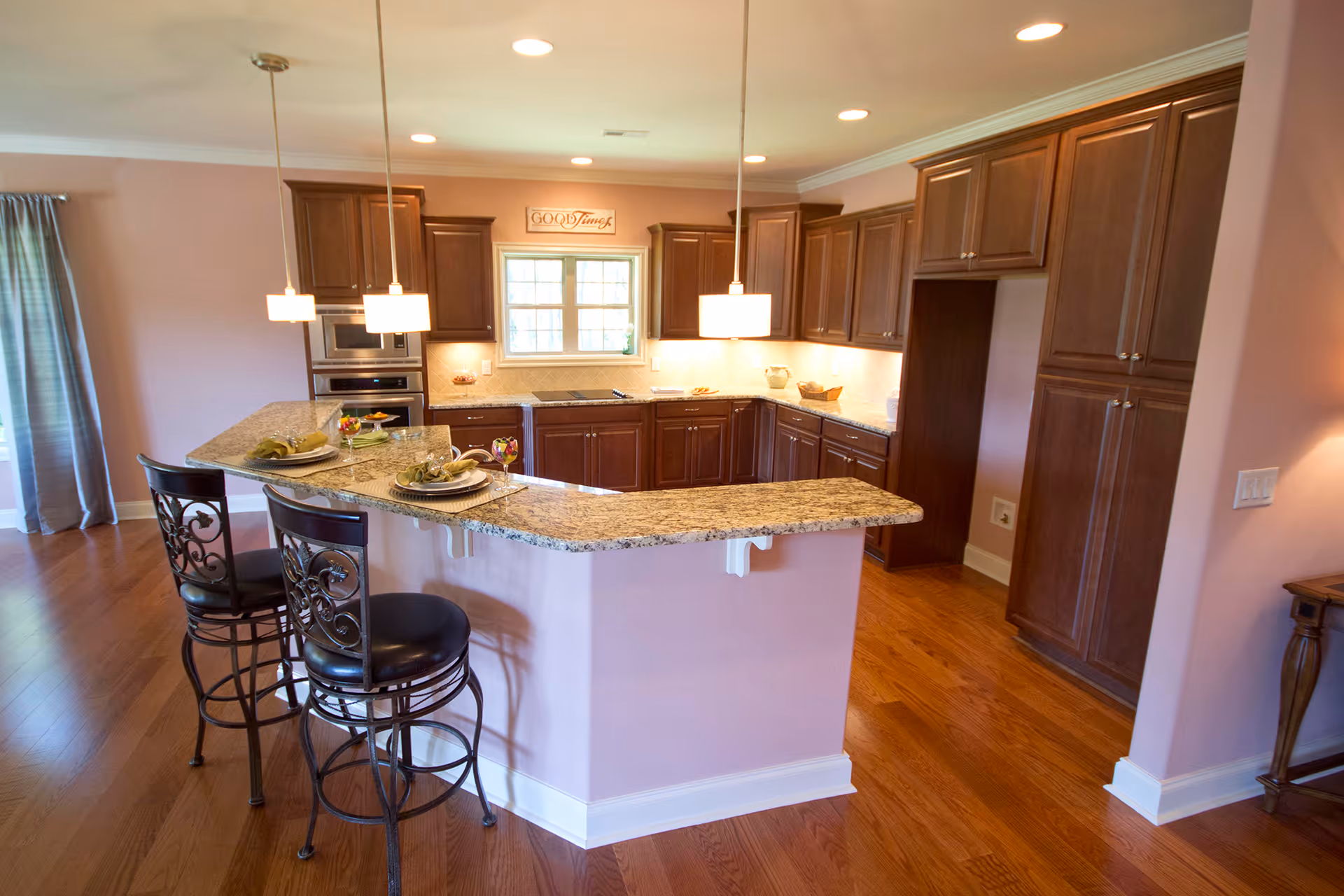 A spacious kitchen with wooden cabinets and a granite countertop island with two high chairs. The kitchen has hardwood floors, pendant lights hanging above the island, and a window above the sink. There is a sign above the window that reads 'Good Times'.