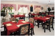 Dining room with multiple tables covered in red tablecloths, each set with placemats and small floral centerpieces. The room has large windows with pink valances, floral upholstered chairs, and a bright, clean atmosphere.