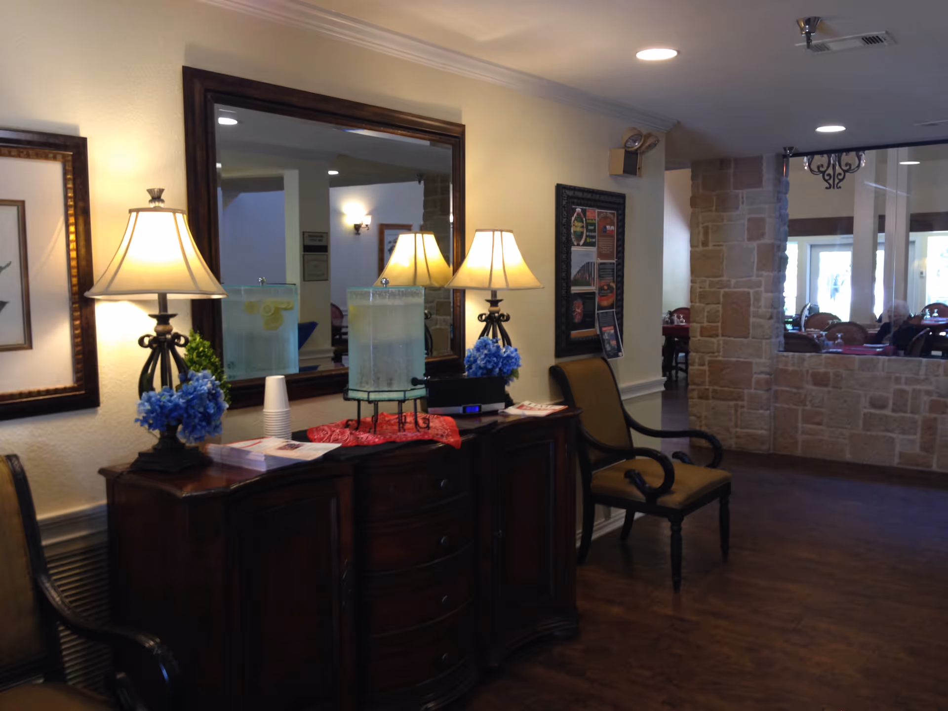 Interior common area with a wooden console table topped with lamps, blue flowers, and drink dispensers, with chairs and a dining area visible through an opening.
