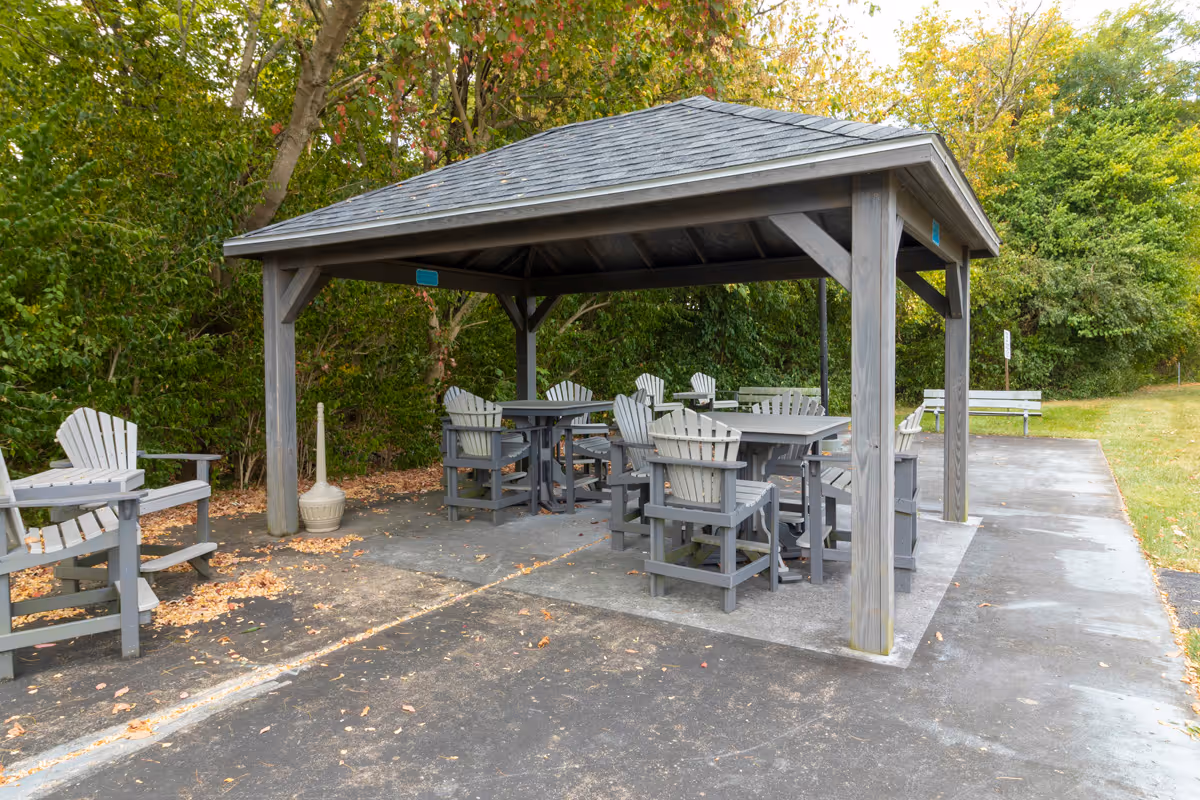 Outdoor seating area with a wooden gazebo covering several gray Adirondack chairs and tables, surrounded by trees and greenery.