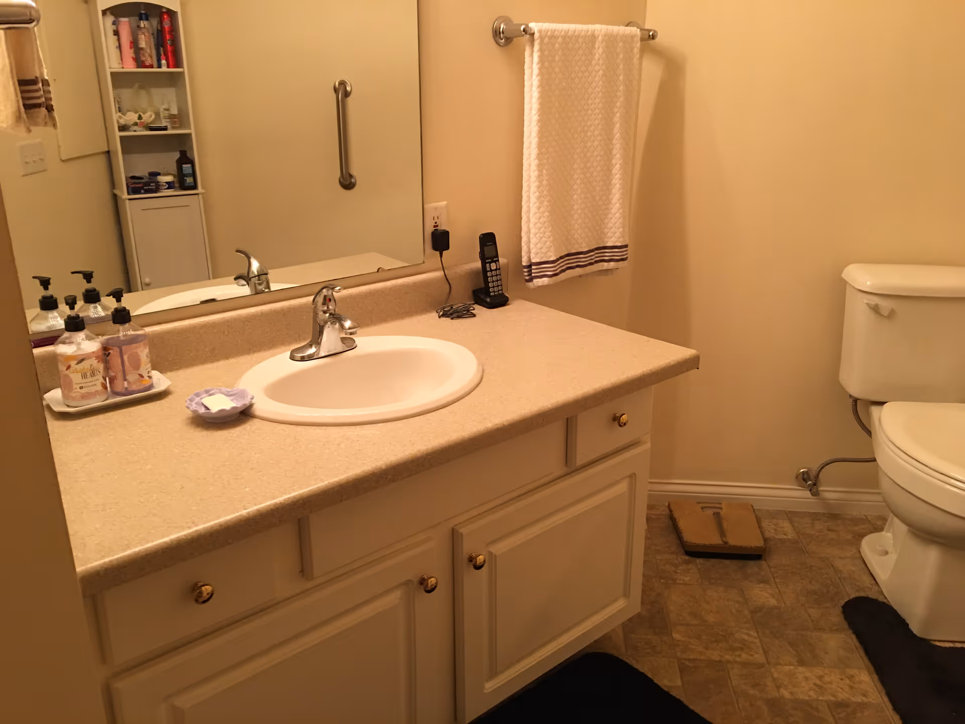 A bathroom with a beige countertop and white sink. On the countertop are three soap dispensers and a small dish with a bar of soap. A large mirror is mounted above the sink. To the right, there is a white toilet and a towel hanging on a silver towel rack. The floor has a patterned tile, and there is a black rug in front of the sink and toilet. A small shelving unit with toiletries is visible in the mirror's reflection.