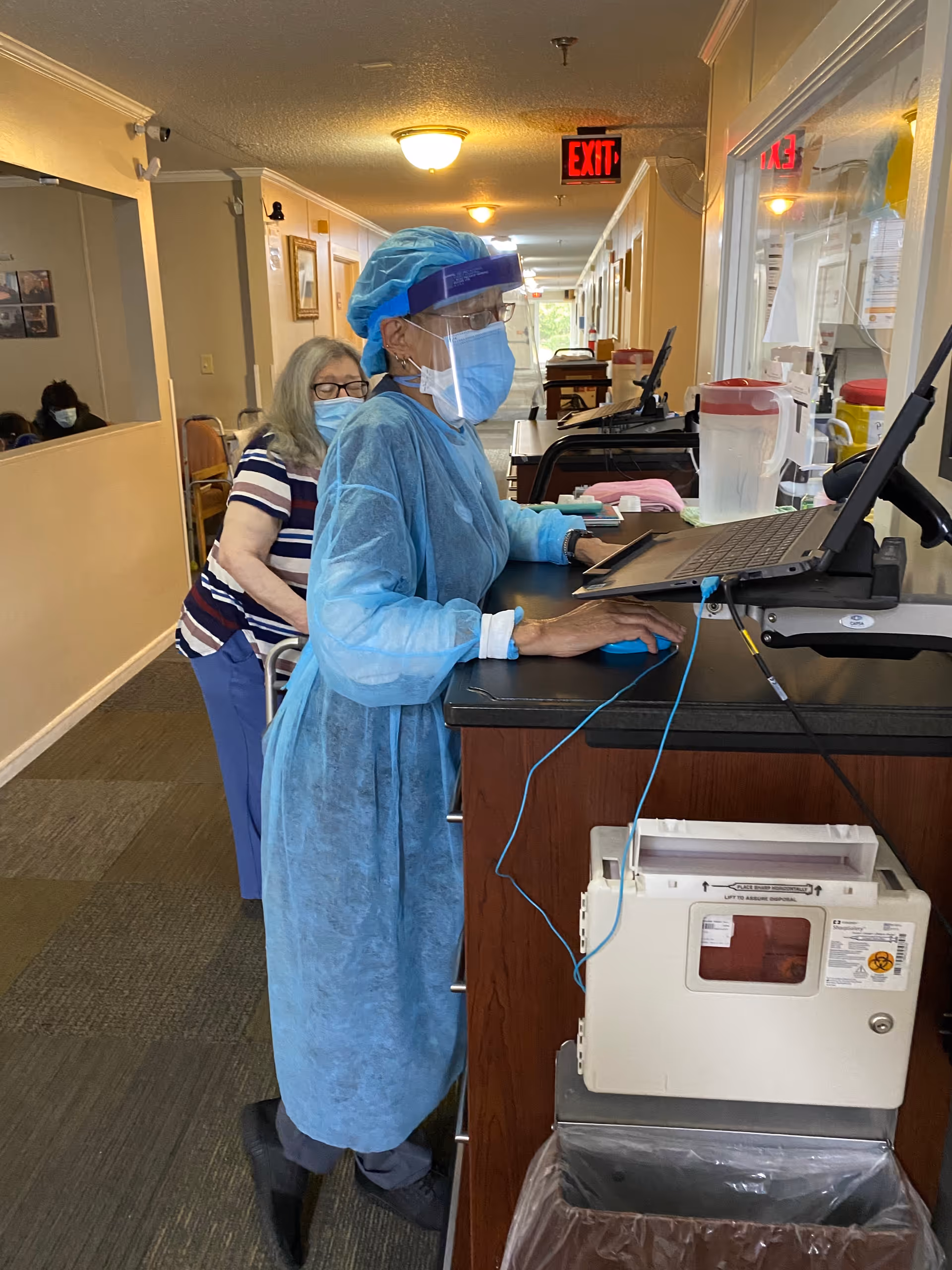A healthcare worker wearing full protective gear including a blue gown, face mask, face shield, and hair cover is standing at a counter using a laptop in a hallway. Behind the worker, an elderly woman wearing a mask is standing with a walker. The hallway has beige walls, carpeted floors, and an exit sign visible in the background.