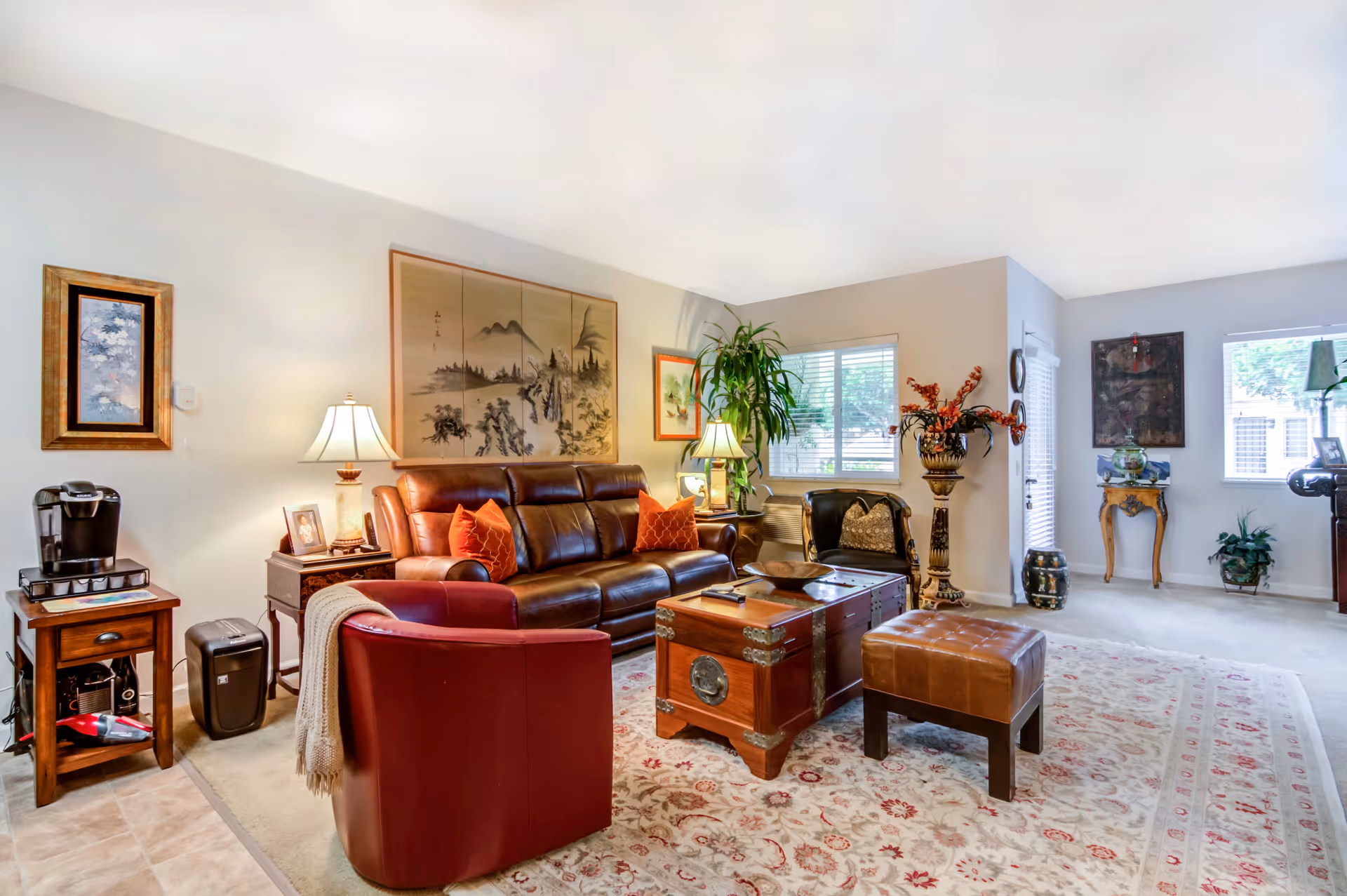 A bright and cozy living room with a leather sofa, a red armchair, a wooden chest used as a coffee table, and a leather ottoman. The room features a large patterned area rug, several plants, framed artwork on the walls, and two table lamps providing warm lighting. There is a coffee maker on a small wooden side table near the entrance to the room.