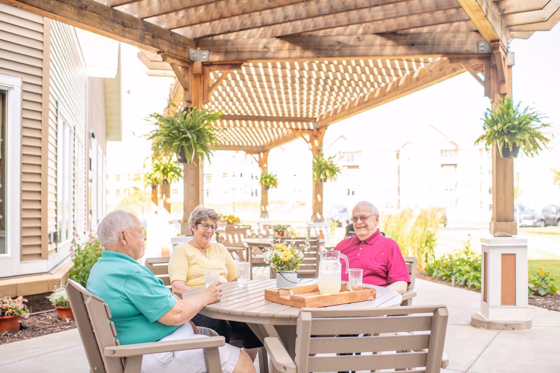 Three elderly people sitting around an outdoor table under a wooden pergola enjoying drinks and conversation.