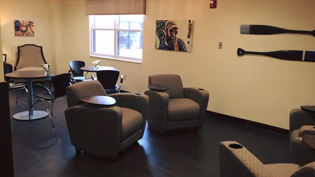 A cozy interior room with several cushioned armchairs, small round tables, and a window letting in natural light. The walls are decorated with two paintings and two wooden oars mounted horizontally.
