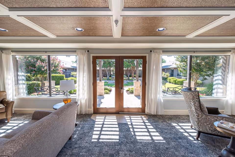 A bright and inviting living room area with large windows on either side of a wooden double door leading outside. The room features a patterned carpet, a sofa, an armchair, a side table with a lamp, and sheer white curtains. Outside, there is a well-maintained garden with trees and shrubs.