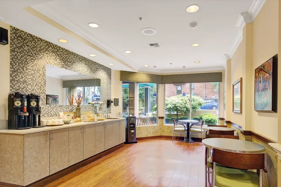 A bright dining area in a senior living facility with wooden floors, beige walls, and large windows showing greenery outside. There are small round tables with chairs, a counter with coffee dispensers, cups, and snacks, and a large mirror with a mosaic tile backsplash above the counter.