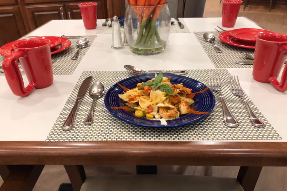 A dining table set with a blue plate of pasta garnished with herbs, surrounded by silverware including knives, forks, and spoons. The table has a white tablecloth with woven placemats, red mugs, and a glass vase with orange tulips in the center.