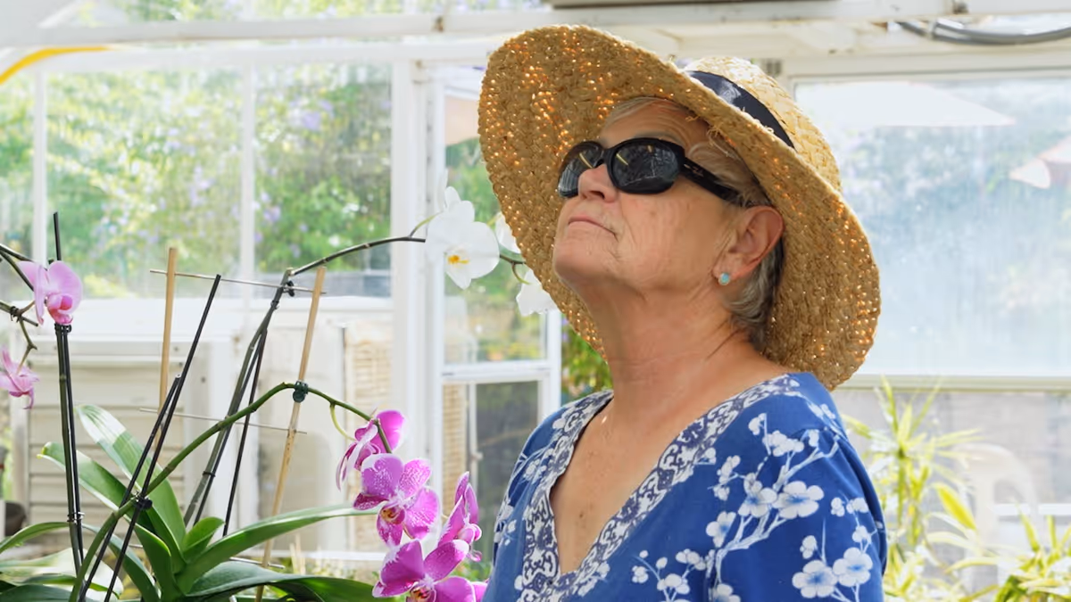 An elderly woman wearing a large straw hat, dark sunglasses, and a blue floral blouse is standing indoors near blooming orchids, looking upwards. The background shows a bright room with large windows and some greenery outside.