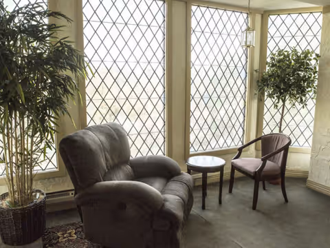 A cozy sitting area with a large cushioned armchair and a wooden chair with armrests, separated by a small round table. The space is illuminated by natural light coming through three tall windows with diamond-patterned glass. There are two potted plants on either side of the seating area.
