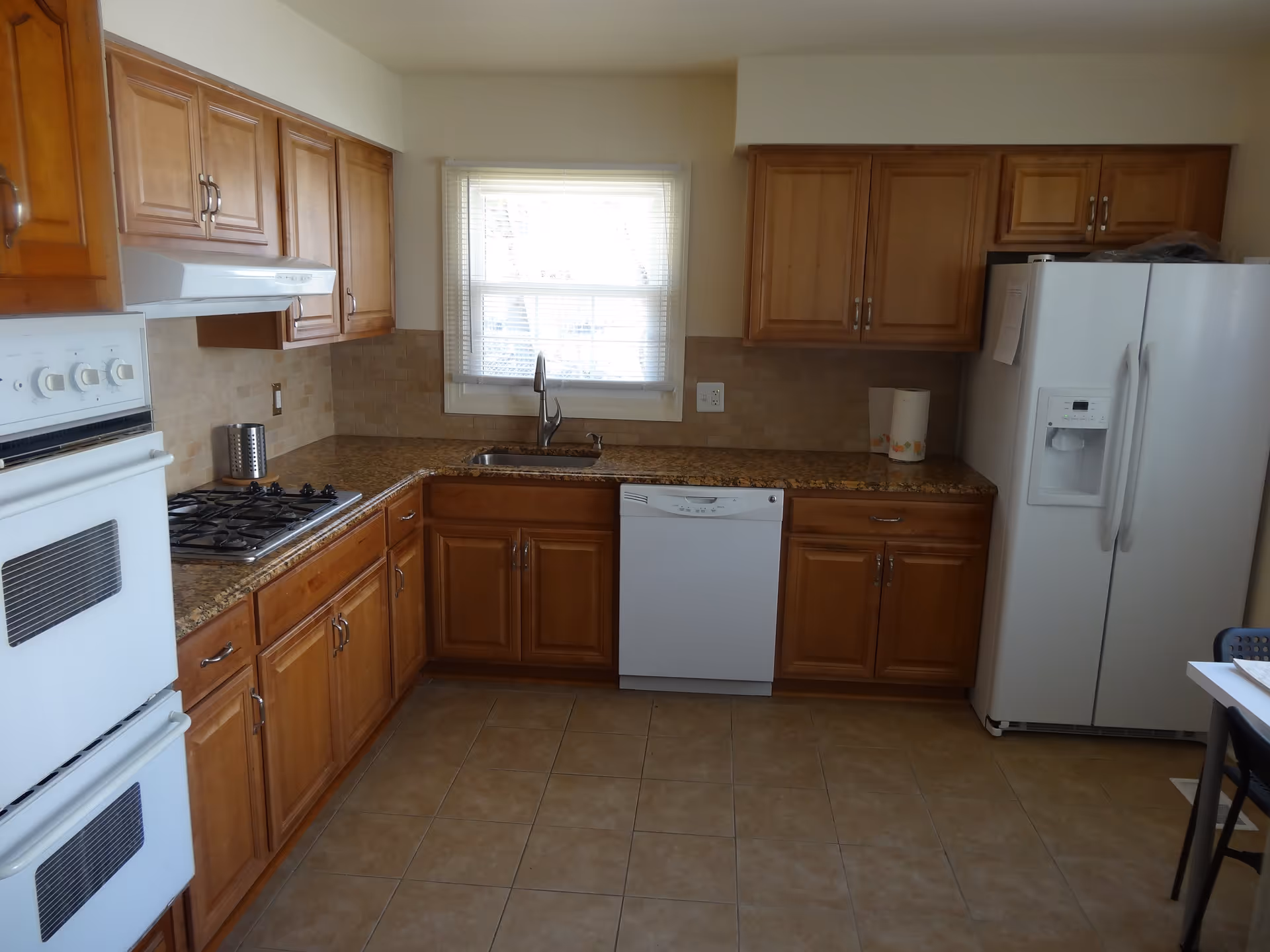 A kitchen with wooden cabinets, a white double oven, a gas stove, a dishwasher, a double-door refrigerator, a sink under a window with blinds, and a tiled floor.