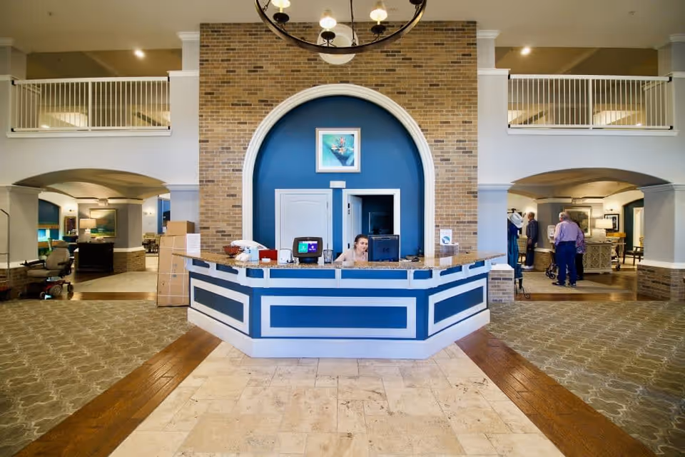 Reception area of Isle at Raider Ranch featuring a central desk with a staff member working on a computer. The area has a brick accent wall with a blue inset and an arched design. There are open spaces on either side with seating and people visible in the background. The floor has a combination of tile, wood, and carpet.