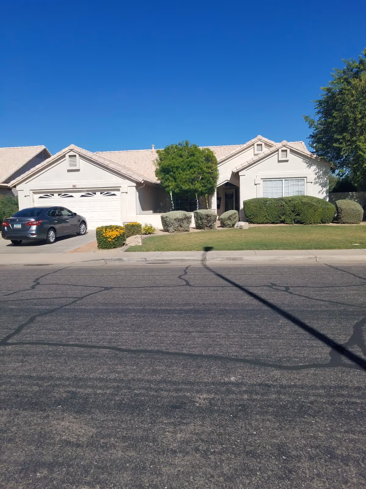 Single-story residential house with a beige exterior, a two-car garage, a driveway with a parked gray car, a well-maintained front lawn with bushes and a tree, under a clear blue sky.