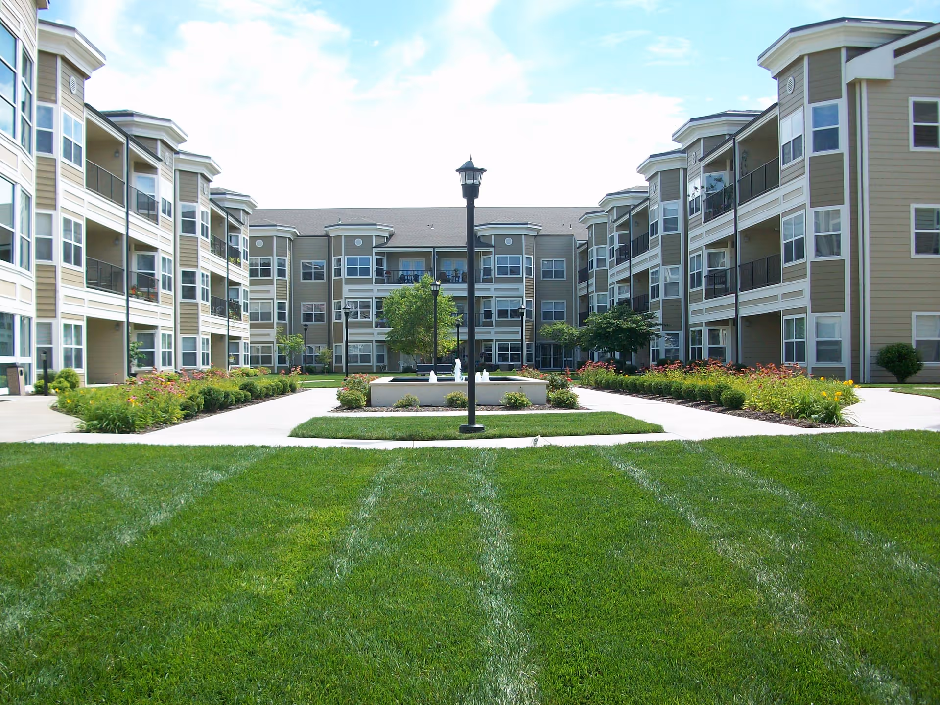 Courtyard view of a senior living complex with a landscaped lawn, central fountain and lamp post, and surrounding three-story buildings with balconies.