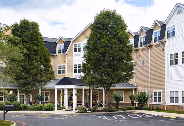 Exterior view of AVIVA Baldwin Park senior living facility showing a multi-story building with beige and white siding, several windows, a covered entrance with white pillars, and landscaped greenery including trees and bushes in front. There is a parking area with marked spaces in the foreground.