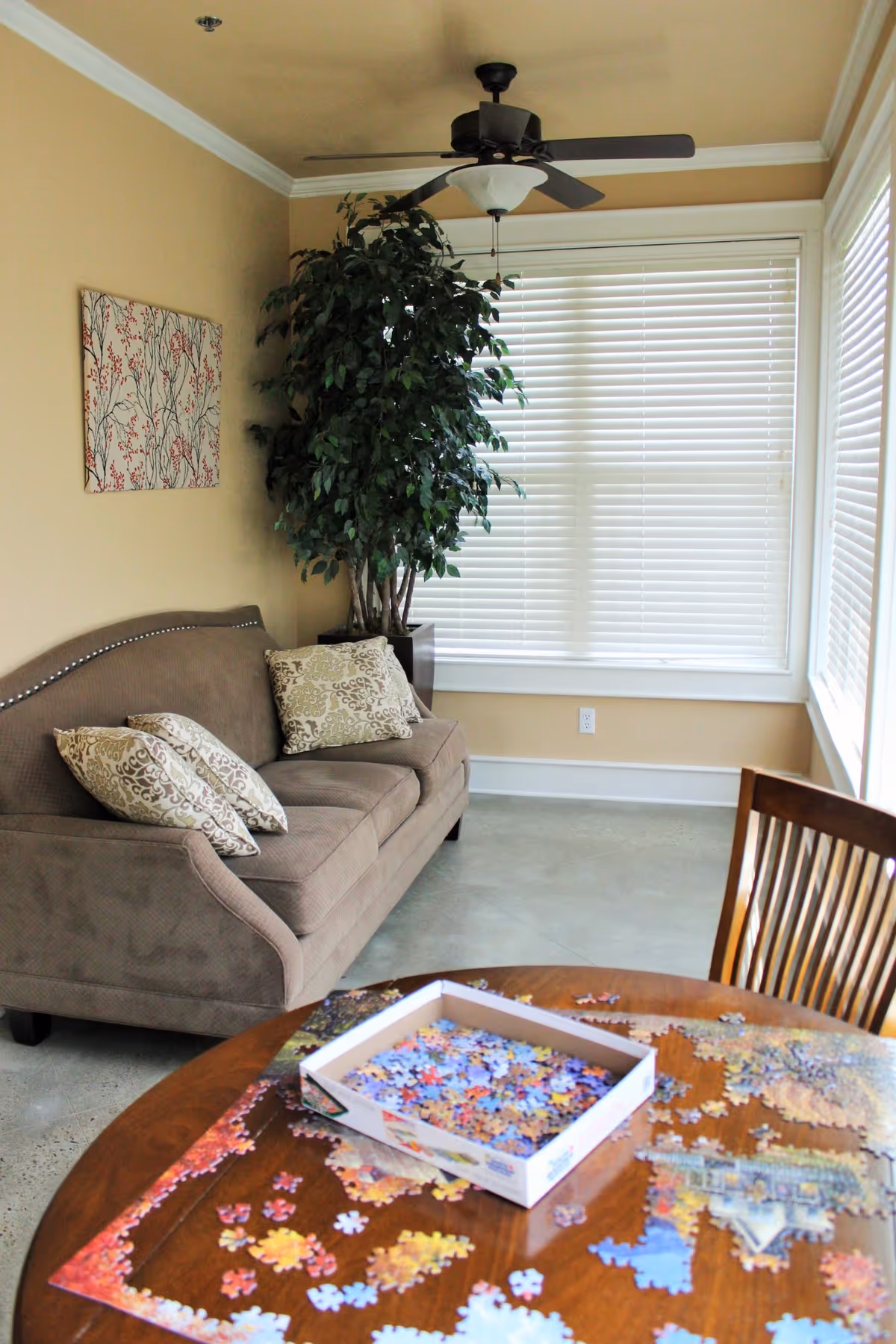 A cozy sitting area with a brown upholstered sofa adorned with patterned cushions, a large potted plant in the corner, and a ceiling fan above. In the foreground, a wooden table holds a partially completed jigsaw puzzle with a box of puzzle pieces.