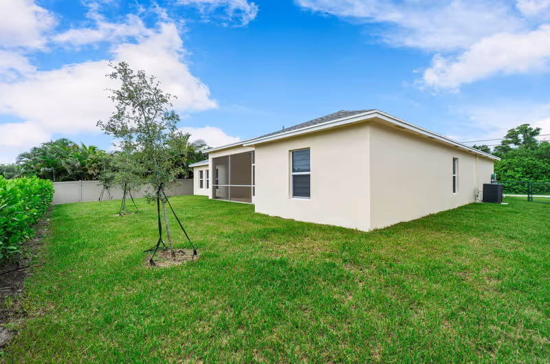 Backyard view of a single-story beige house with a screened porch, green lawn, young trees and a white fence under a blue sky.