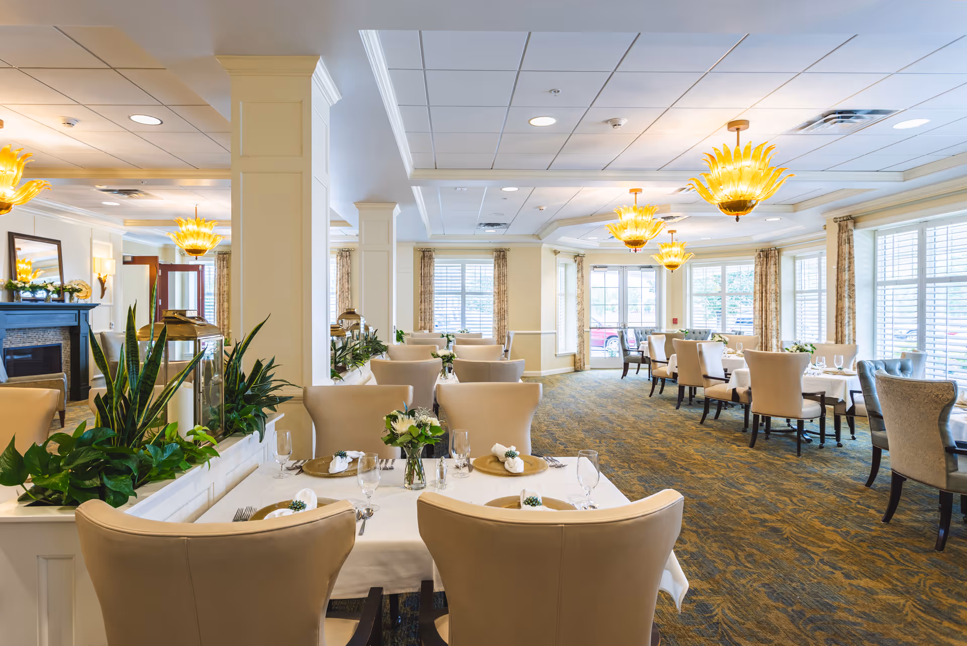 Bright elegant dining room with multiple set tables, beige upholstered chairs, large windows, plants, and distinctive yellow chandeliers.