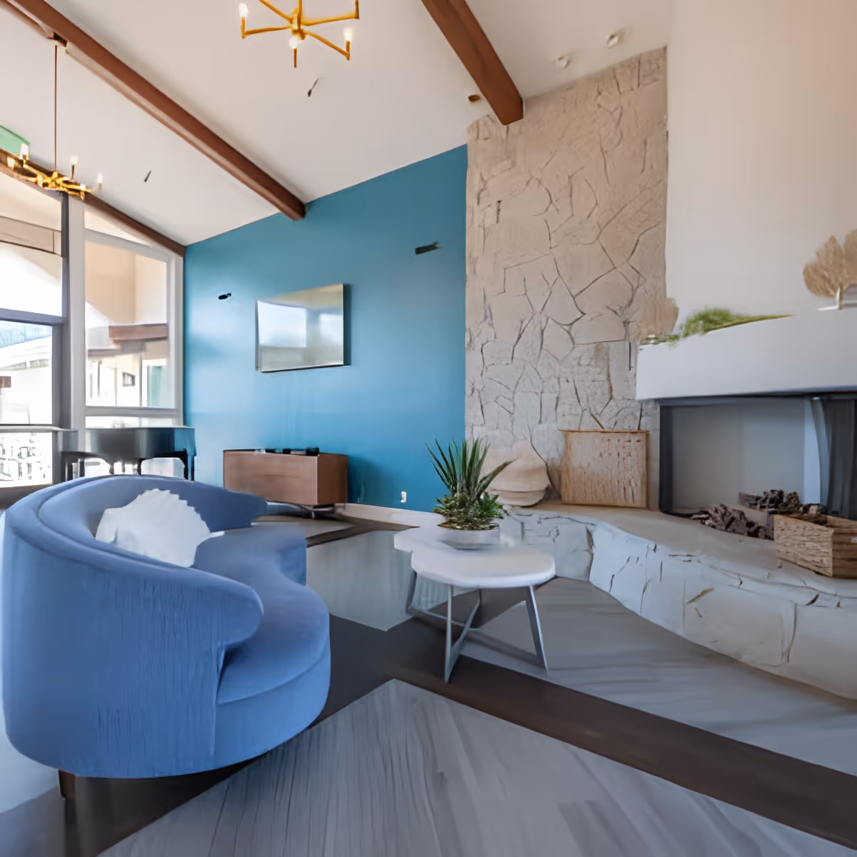 A modern living room with a curved blue sofa, a white round coffee table with a small plant, a stone fireplace with decorative items, a blue accent wall with a mounted flat-screen TV, large windows letting in natural light, and exposed wooden ceiling beams.