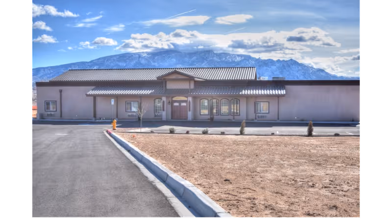 Single-story senior living facility building with a tiled roof and central entrance, a paved parking area and dry landscaping in front with mountains in the background.