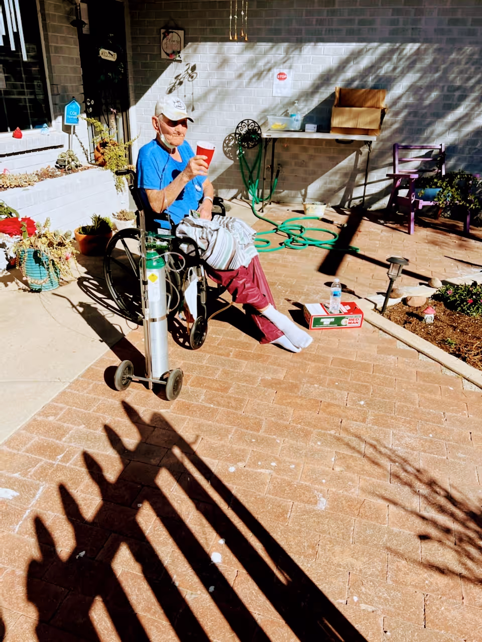 An elderly man in a wheelchair holding a red cup outdoors on a sunny day. He is wearing a blue shirt, maroon pants, white socks, and a white cap. There is an oxygen tank attached to the wheelchair. The setting appears to be a patio area with potted plants, a garden hose, a table with items on it, and a brick wall in the background.