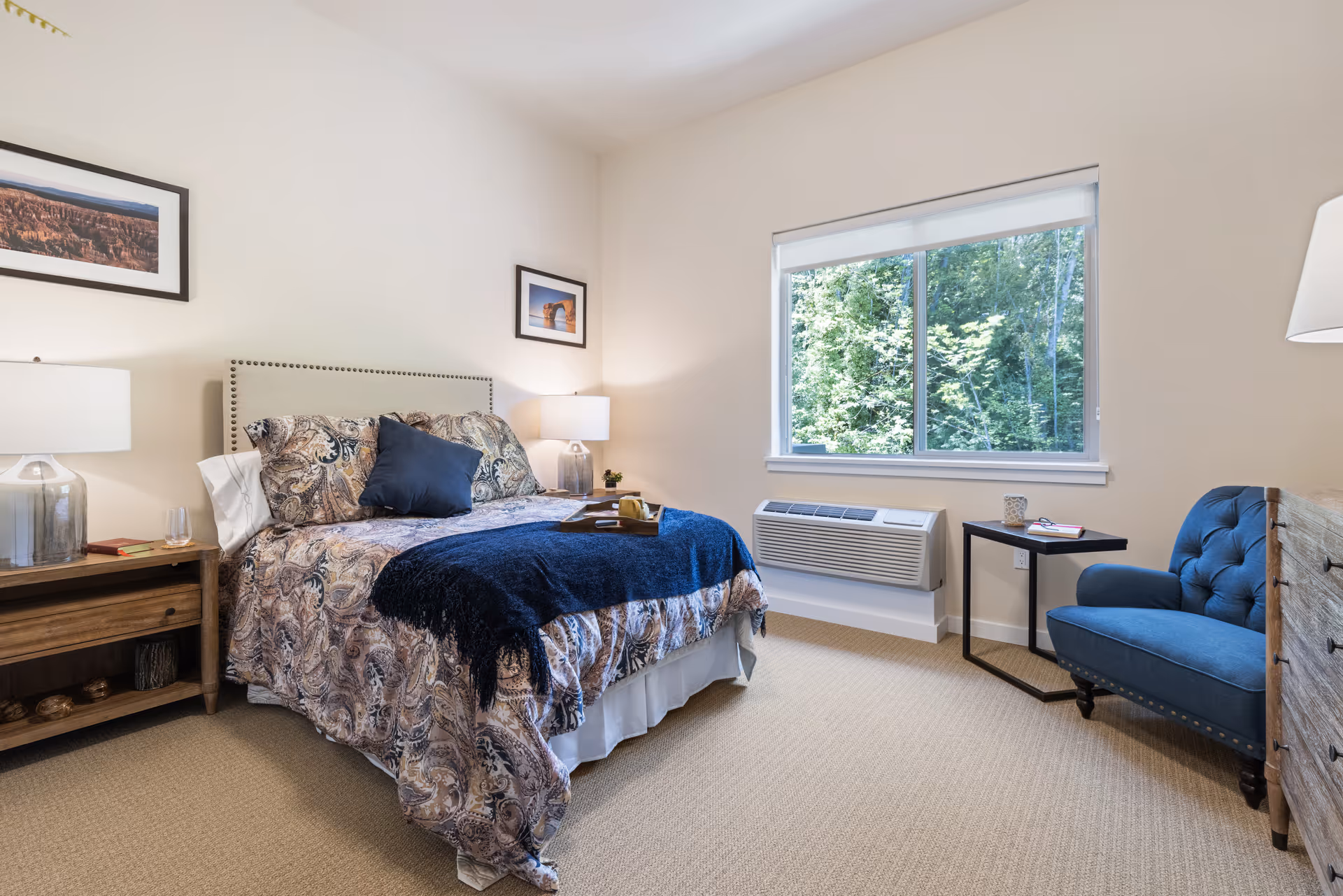 A cozy bedroom with a bed featuring patterned bedding and a navy blue throw blanket. There are two bedside tables with lamps, a blue upholstered chair next to a small black side table, a wooden dresser, and a large window showing green trees outside.