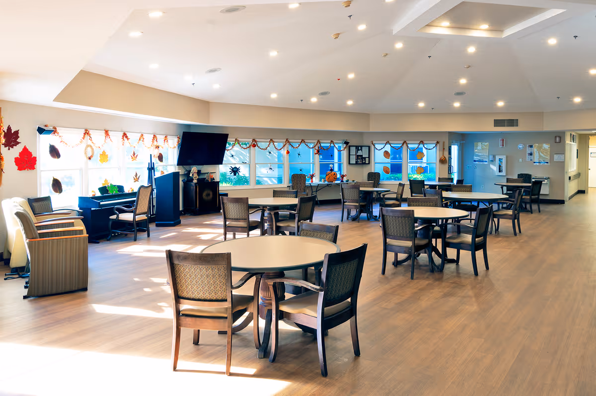 A spacious and well-lit common area with several round tables and chairs arranged for seating. Large windows decorated with autumn-themed decals and garlands let in natural light. There is a television mounted on the wall and a piano near the windows. The room has a high ceiling with recessed lighting and wood flooring.