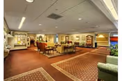 A spacious senior living facility common area with carpeted floors, multiple seating arrangements including sofas and armchairs, framed artwork on the walls, and bulletin boards. The room is well-lit with ceiling lights and has a warm, inviting atmosphere.
